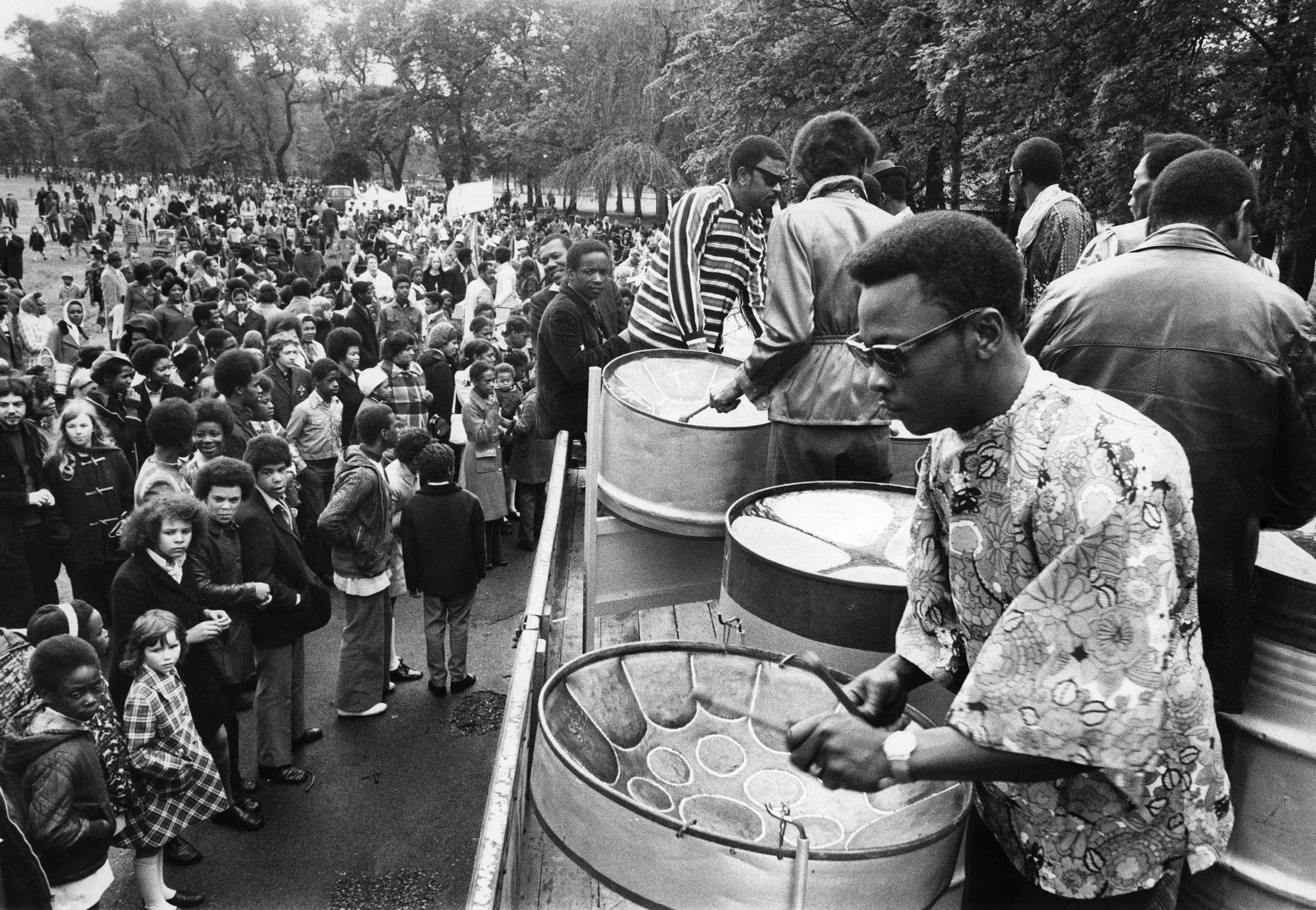 Man with an afro and sunglasses plays a steel drum to a large crowd at the carnival.